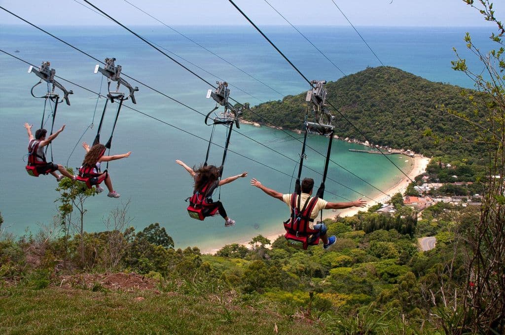 Turistas deslizando em cadeiras de tirolesa do Parque Unipraias com vista para o mar verde-turquesa e a Mata Atlântica ao fundo