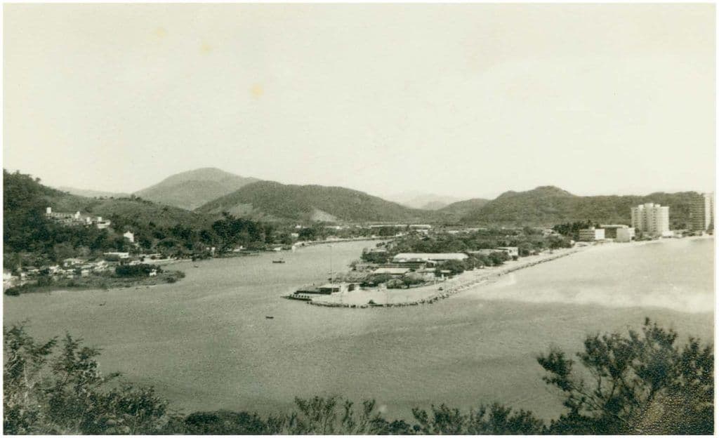 Foto em preto e branco histórica da Barra Sul em Balneário Camboriú, mostrando casas de veraneio à beira da água e um pequeno porto com colinas ao fundo.