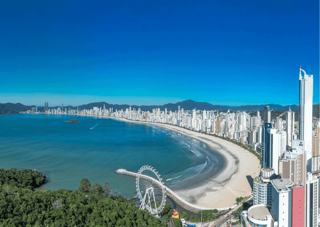 Vista panorâmica aérea da orla de Balneário Camboriú com roda-gigante à beira-mar e prédios altos contornando a curva da praia.