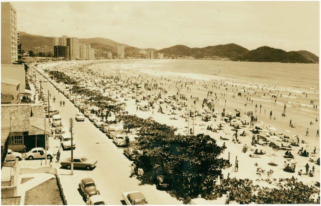 Imagem vintage sépia de uma avenida à beira-mar em Balneário Camboriú, com carros antigos estacionados e dezenas de banhistas espalhados pela faixa de areia.