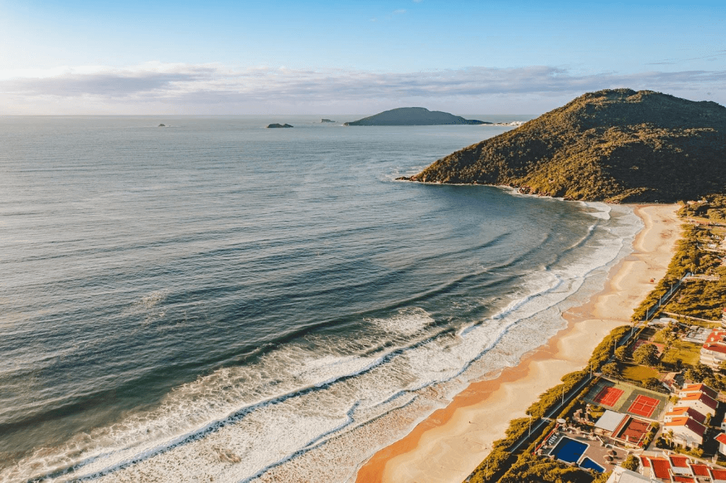 Vista aérea de uma praia em Santa Catarina, mostrando mar azul, montanhas cobertas de vegetação e construções próximas à areia