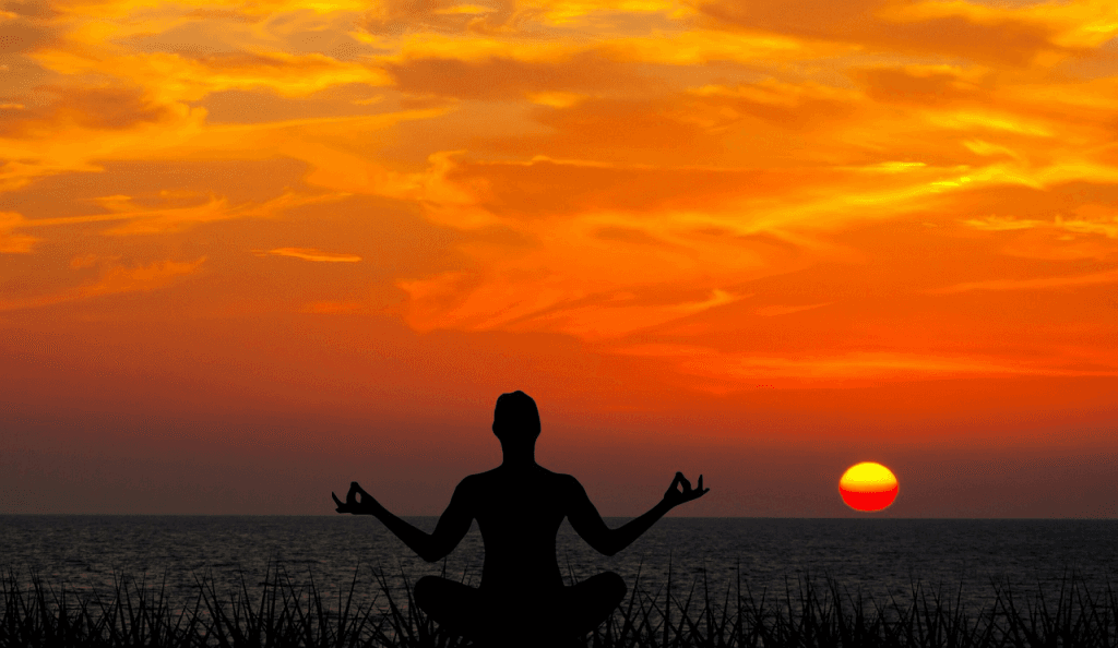Silhueta de pessoa sentada em posição de lótus, braços abertos com as mãos em mudra, diante do mar calmo ao pôr-do-sol, com o céu pintado de tons laranja e dourado.